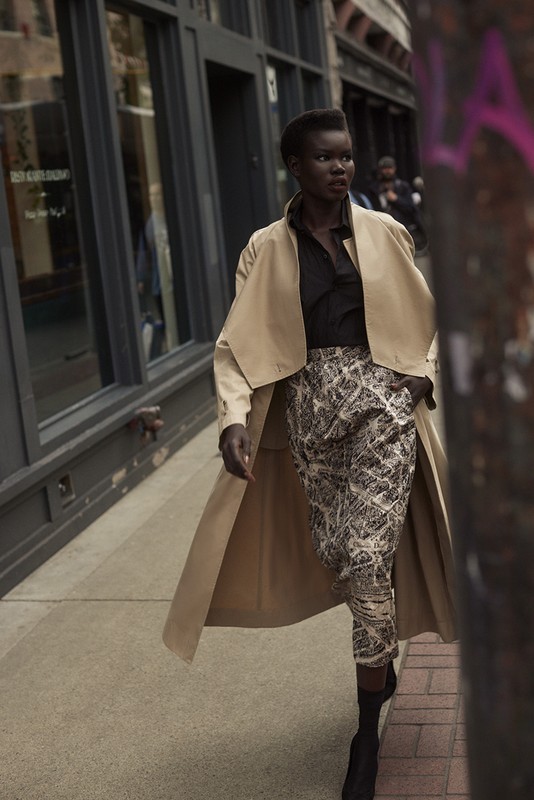 Fashionable woman in a trench coat walks a city sidewalk, wearing patterned pants and boots, posing in an urban street scene.