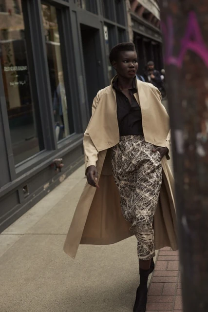Fashionable woman in a trench coat walks a city sidewalk, wearing patterned pants and boots, posing in an urban street scene.