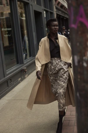 Fashionable woman in a trench coat walks a city sidewalk, wearing patterned pants and boots, posing in an urban street scene.