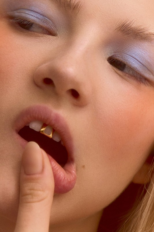 Close-up of a woman's face with blue eyeshadow, finger touching lip, and gold dental jewelry on her front teeth in soft lighting.
