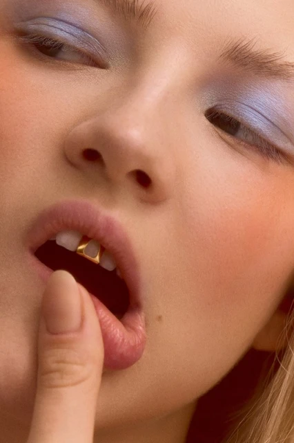 Close-up of a woman's face with blue eyeshadow, finger touching lip, and gold dental jewelry on her front teeth in soft lighting.