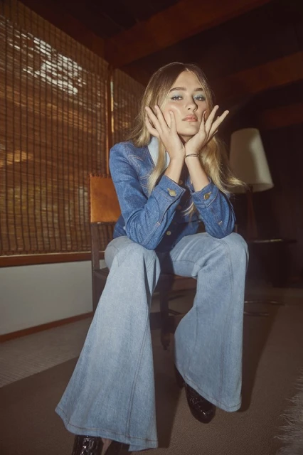 A woman with blonde hair sits in a chair wearing a blue denim jacket and light blue bell-bottom jeans in an indoor setting.