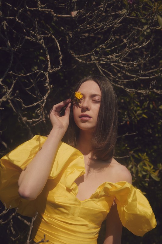 Portrait of a young woman in a yellow dress outside, holding a dandelion flower to her eye in a serene outdoor setting.