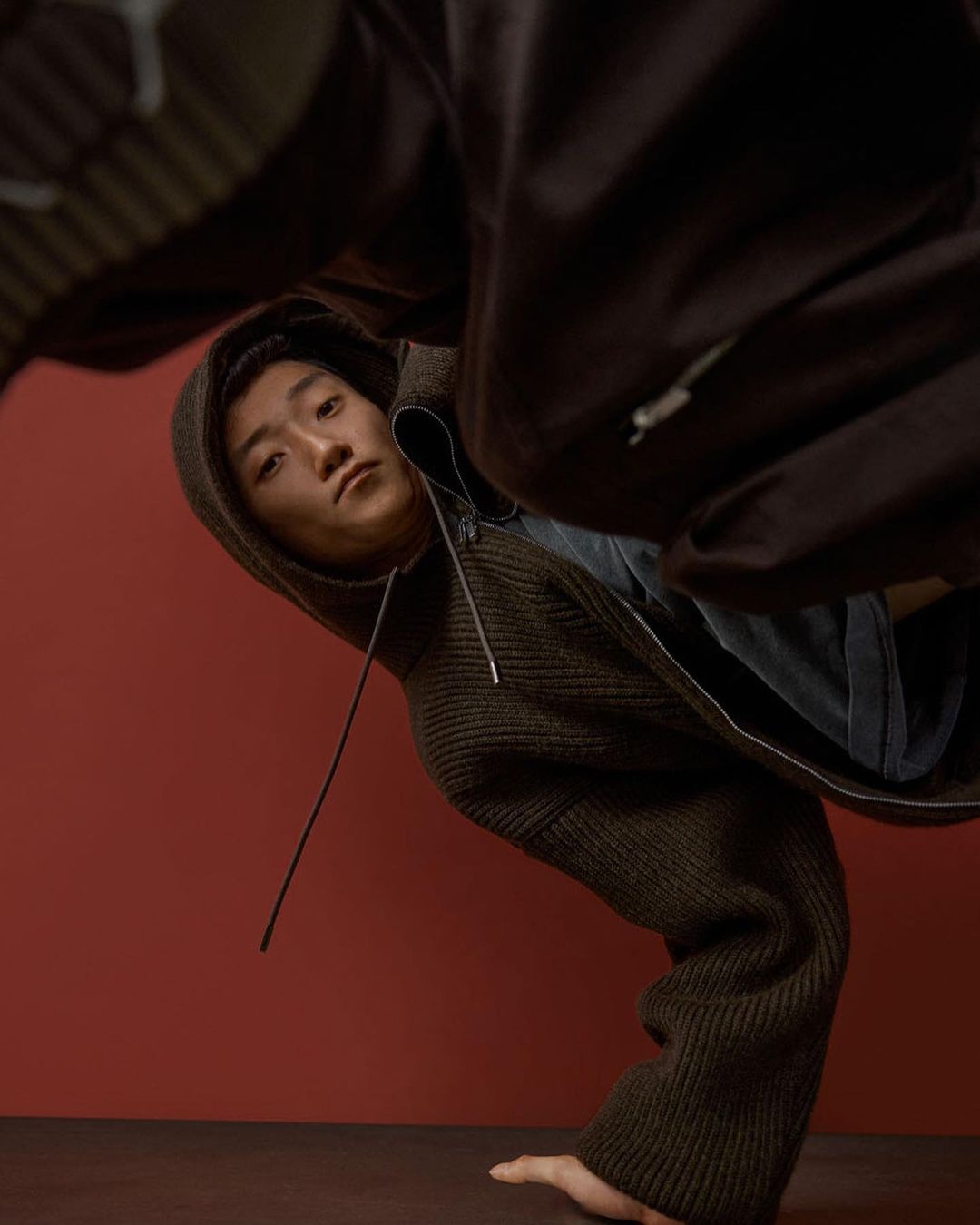 Asian man in hooded jacket strikes a pose with head tilted, angled view against a plain red backdrop. Fashion.
