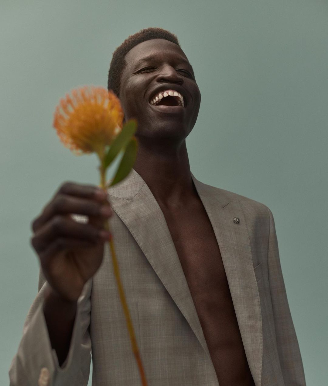 Smiling Black man in a grey suit jacket, with bare chest, holds an orange flower against a pale-blue background in a medium shot.