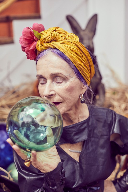Elderly woman with purple hair and a gold turban gazes at a crystal ball in an indoor setting; concrete rabbit statue in background.