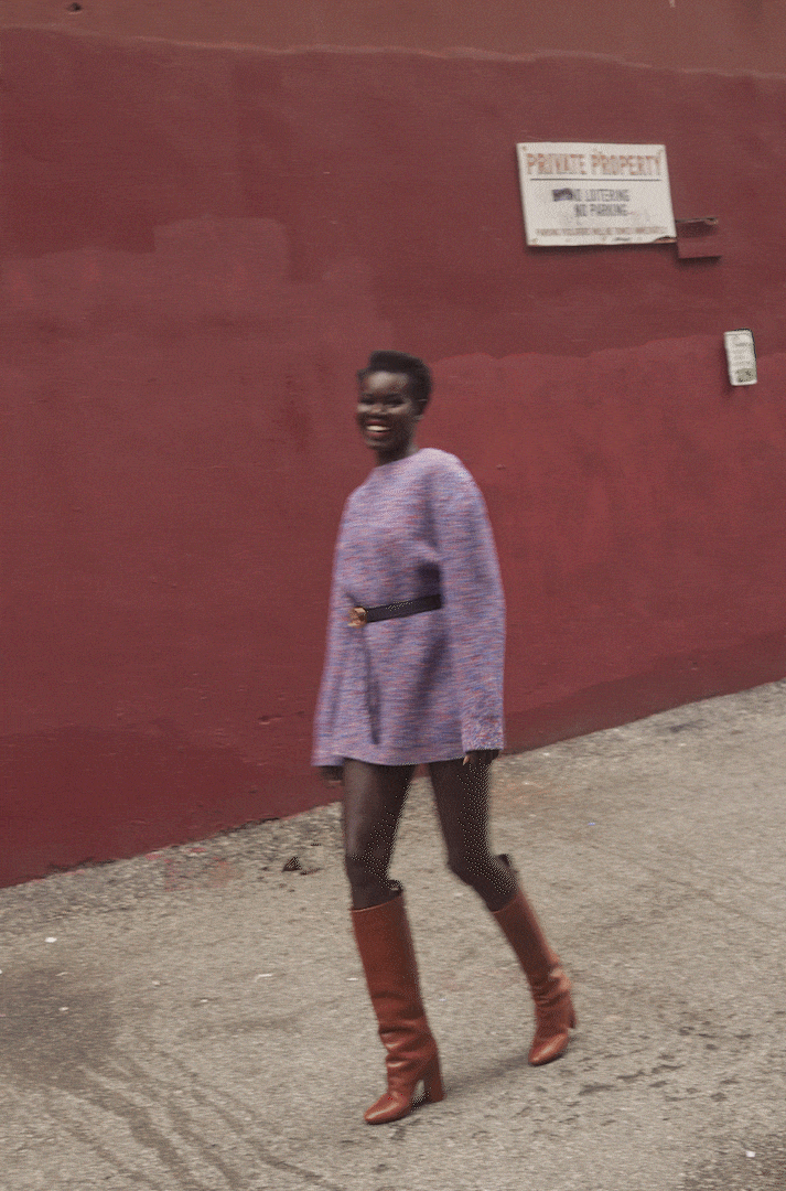 A smiling African-American woman confidently struts down a city sidewalk in a lilac sweater dress and brown knee-high boots.