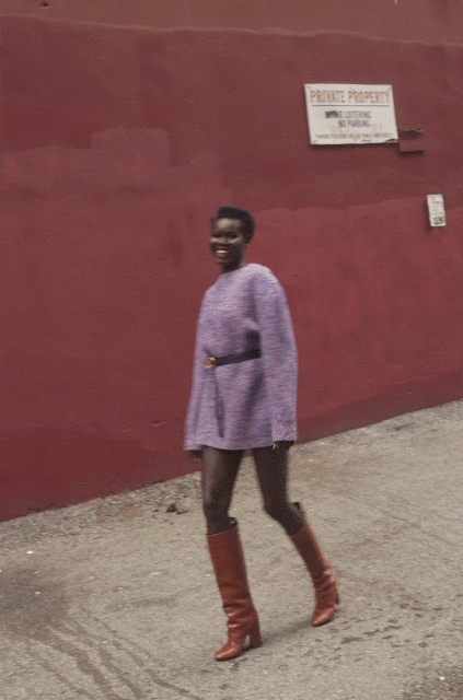 A smiling African-American woman confidently struts down a city sidewalk in a lilac sweater dress and brown knee-high boots.