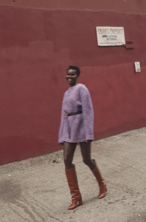 A smiling African-American woman confidently struts down a city sidewalk in a lilac sweater dress and brown knee-high boots.