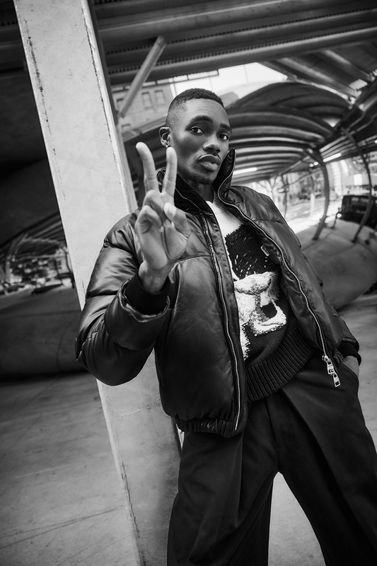 Portrait of a Black male fashion model in a puffer jacket giving a peace sign while leaning against a concrete support in an urban setting.