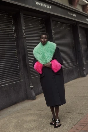 Black woman wearing a long black coat with bright green fur collar and hot pink fur cuffs standing on city sidewalk.