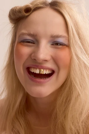 Close-up portrait of a smiling blonde woman with gold teeth and blue eyeshadow in a studio setting. The image has a beauty and fashion aesthetic.