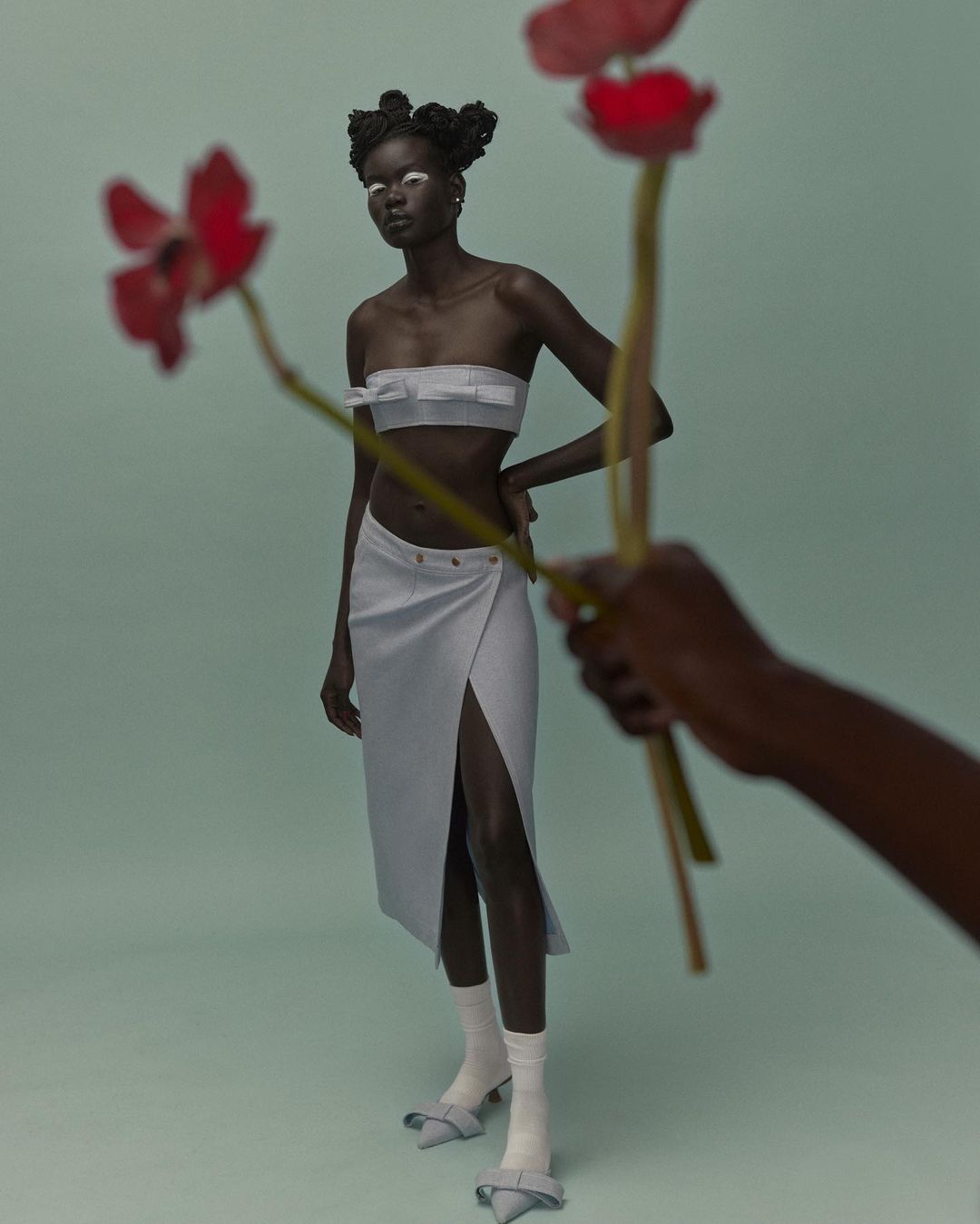 Fashion shoot of a Black female model in a two-piece skirt set posing with red flowers held by a partial view of another person's arm.