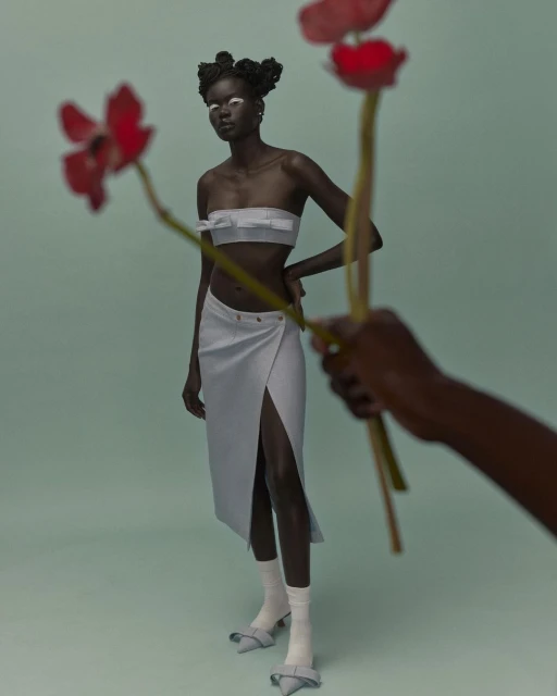 Fashion shoot of a Black female model in a two-piece skirt set posing with red flowers held by a partial view of another person's arm.