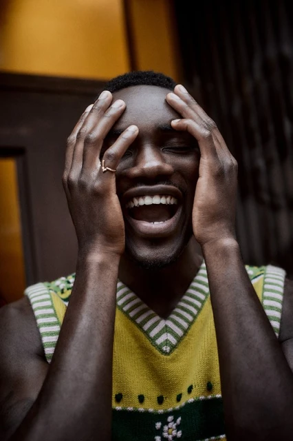 Portrait of an adult African-American male with short hair, wearing a patterned sweater, with his hands on his head and a happy facial expression.