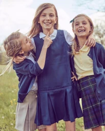 Three young girls smiling together outdoors, wearing school uniforms including navy blue jumpers, cardigans, and a plaid skirt.