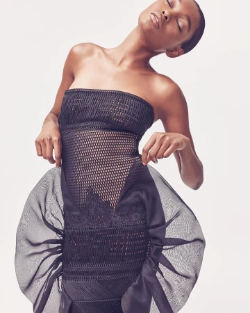 Model wearing a structured black strapless dress with sheer flared panniers against a clean white background.
