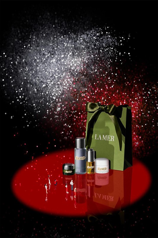 La Mer skincare products including cream, serum, and lotion, are grouped in front of a gift bag against a dark, glittery background.