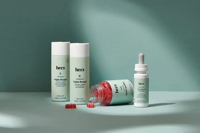 Hers hair products including shampoo, conditioner, biotin builder bottle, and gummy supplements arranged on a two-tone background.