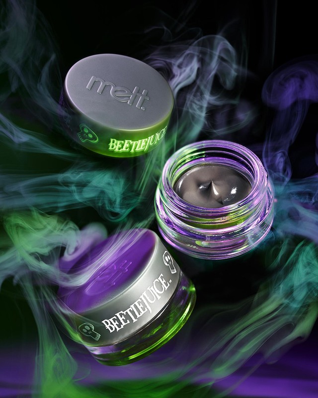 Three 'Beetlejuice' branded cosmetics containers sit against a smoky, colorful background in a studio product shot for beauty advertising.