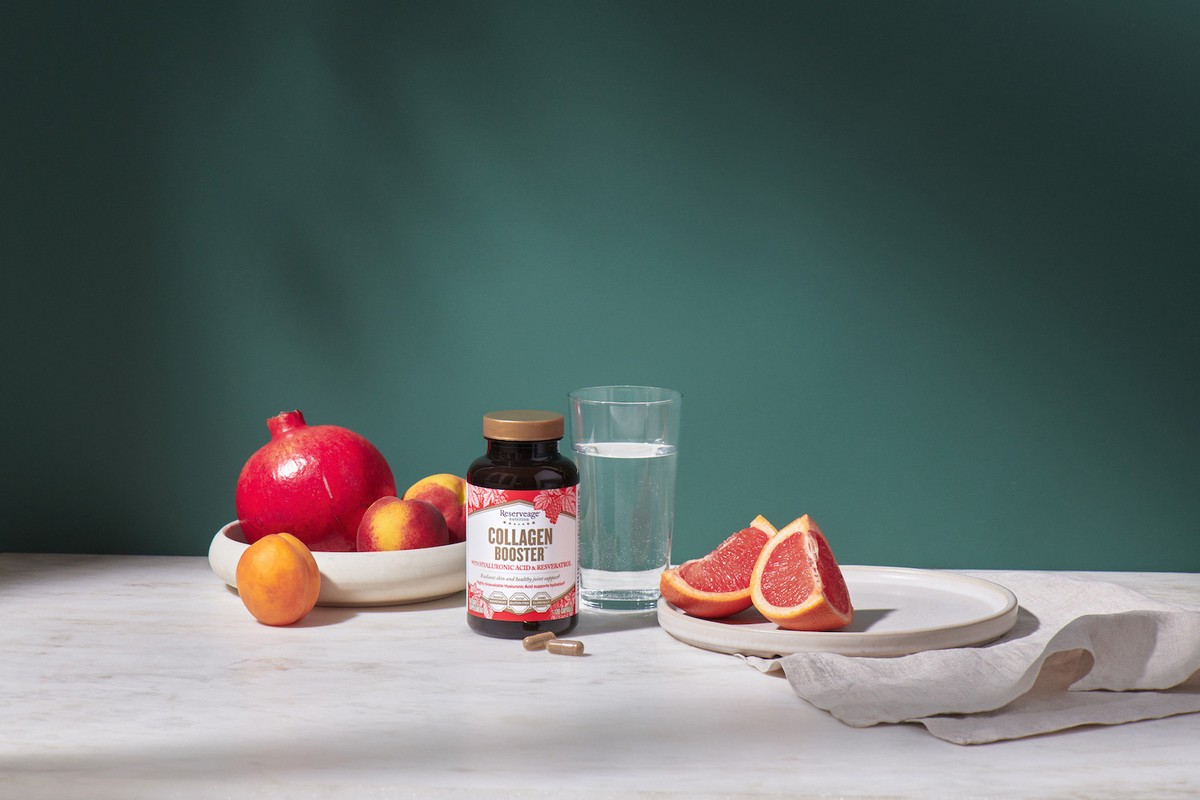 Collagen supplement bottle on a white table with fresh fruits, a glass of water, and a green background, healthy lifestyle.