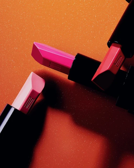 Close-up of three lipsticks in varying shades of pink and coral, arranged against a sparkling orange background, beauty and cosmetics.