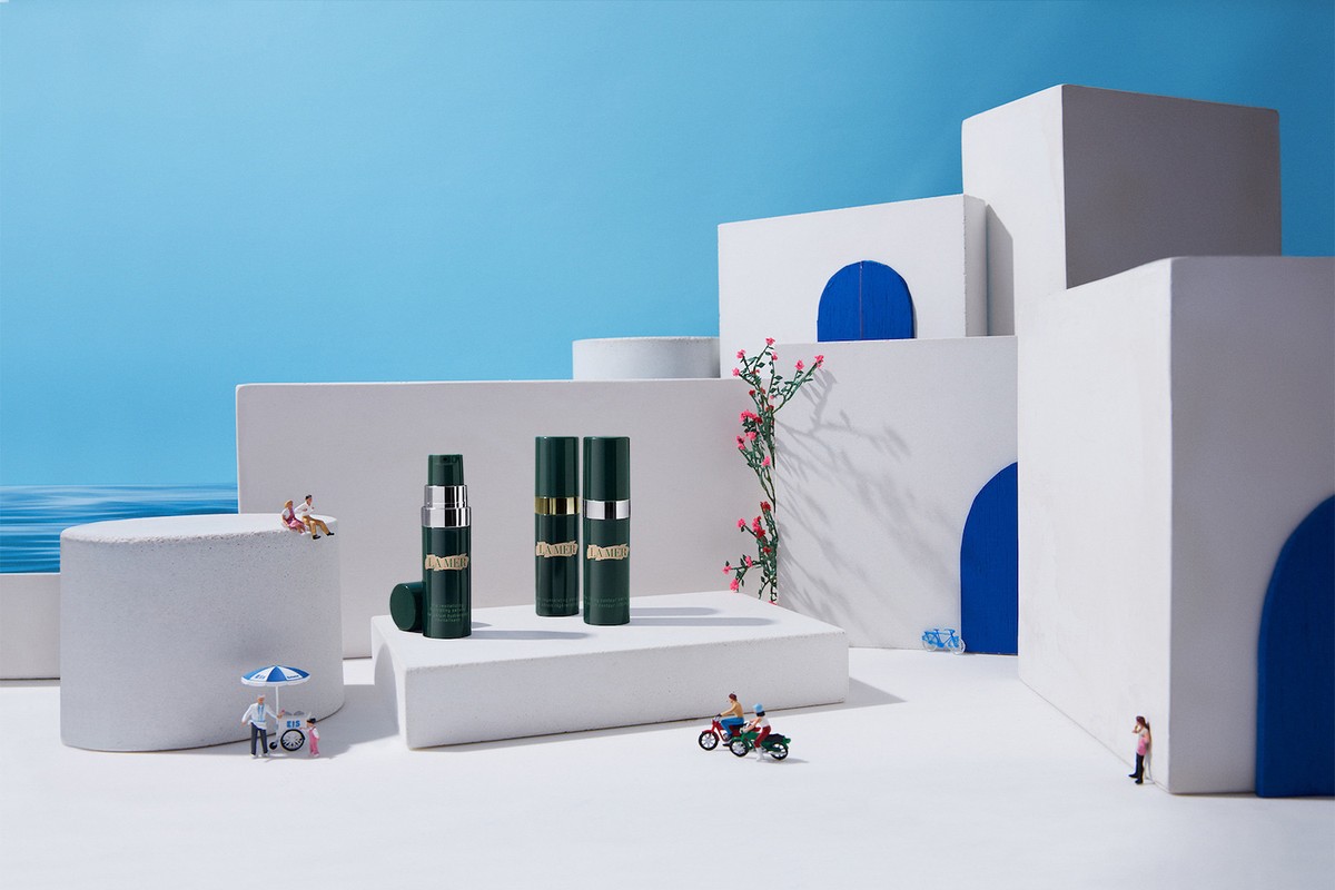 Still life of three La Mer skincare bottles on white platforms, with miniature people and architecture against a blue backdrop.
