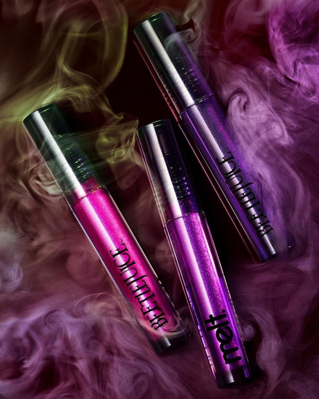 Close-up studio shot of three metallic lip gloss tubes in shades of pink and purple, arranged on a smoky, color-gradient background.