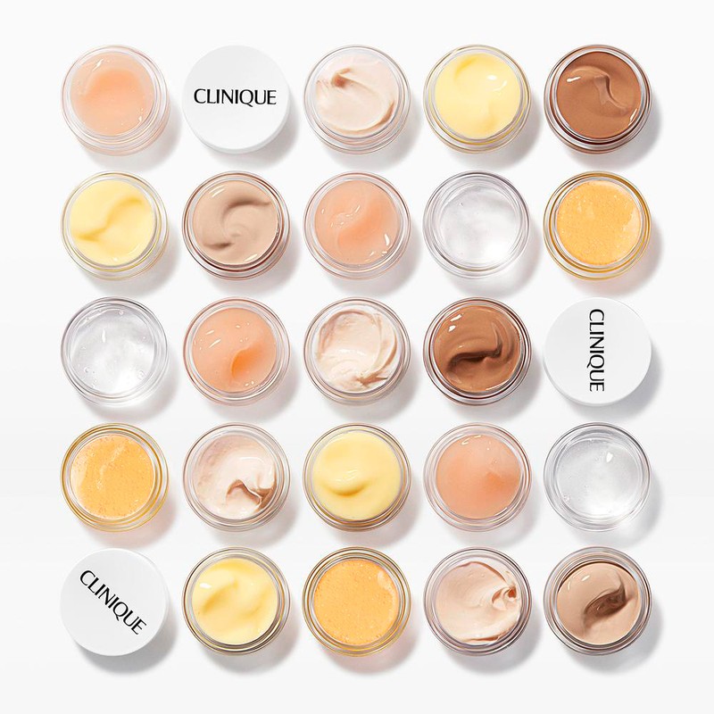 Overhead shot of multiple small clear jars filled with various shades of Clinique face cream, arranged in a grid on a white surface.