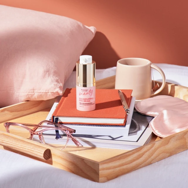 Beauty product on a wooden tray with a book, sleep mask, glasses, and a mug, all on a bed with pink and coral color palette.