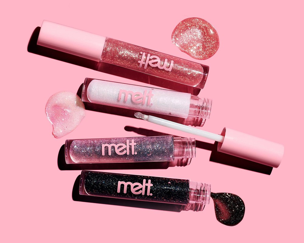 Four Melt lip gloss tubes arranged horizontally on a pink surface with glossy liquid product swatches and an applicator wand.