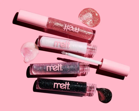 Four Melt lip gloss tubes arranged horizontally on a pink surface with glossy liquid product swatches and an applicator wand.