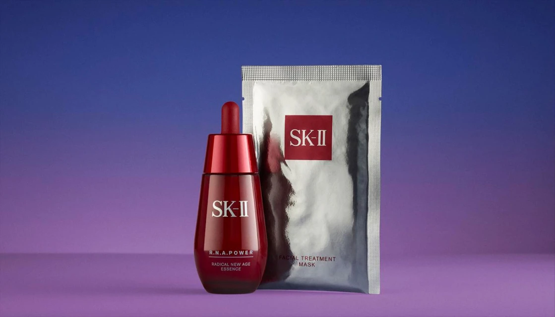 Still life shot of SK-II R.N.A. Power Radical New Age Essence in a red bottle, alongside a silver SK-II Facial Treatment Mask packet.