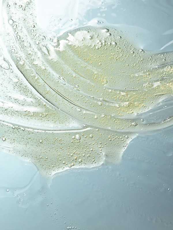 Close-up of viscous, transparent gel with small bubbles, flowing lines, and pale yellow tint against a light blue background. 