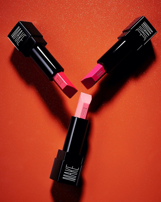 Three black square lipstick tubes with exposed pink and peach bullets arranged in a triangular formation on a textured orange background.
