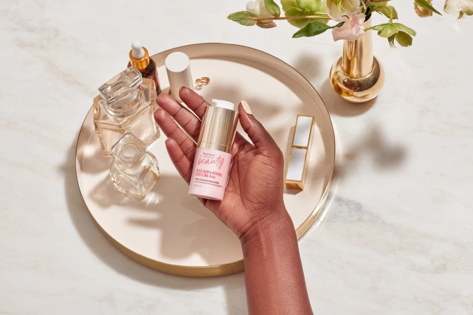 Close-up of a hand holding beauty cream, surrounded by cosmetics, perfume bottles, and flowers on a marble surface in a beauty setting.