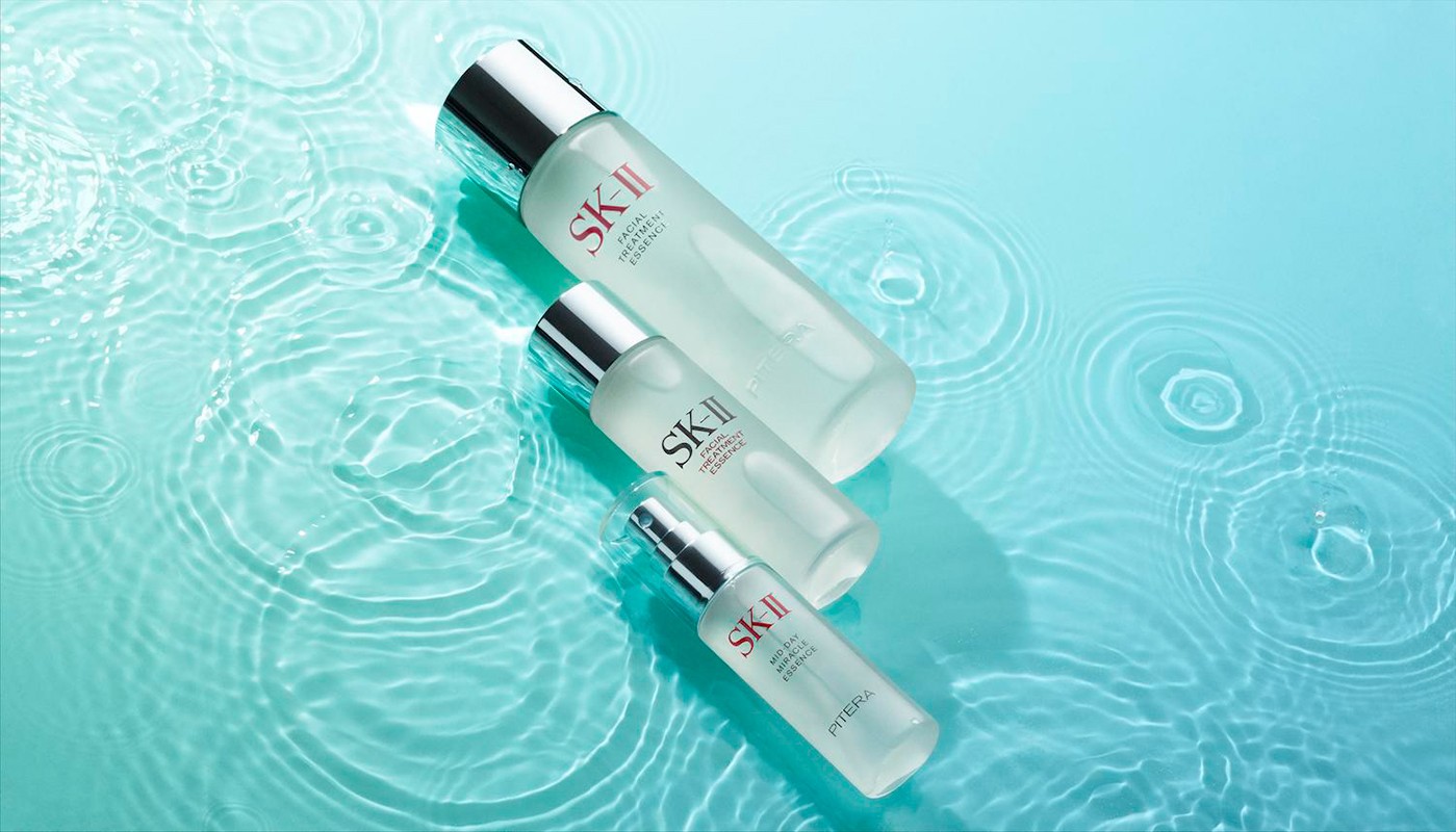Three frosted glass bottles of SK-II facial essence are displayed on a light blue surface with simulated water ripple patterns.