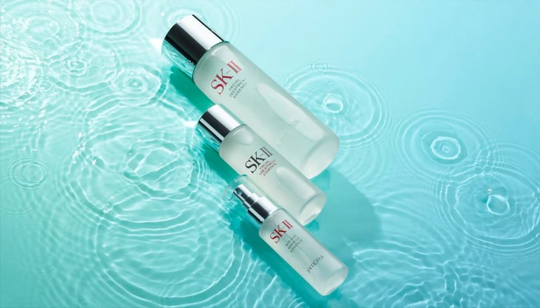 Three frosted glass bottles of SK-II facial essence are displayed on a light blue surface with simulated water ripple patterns.