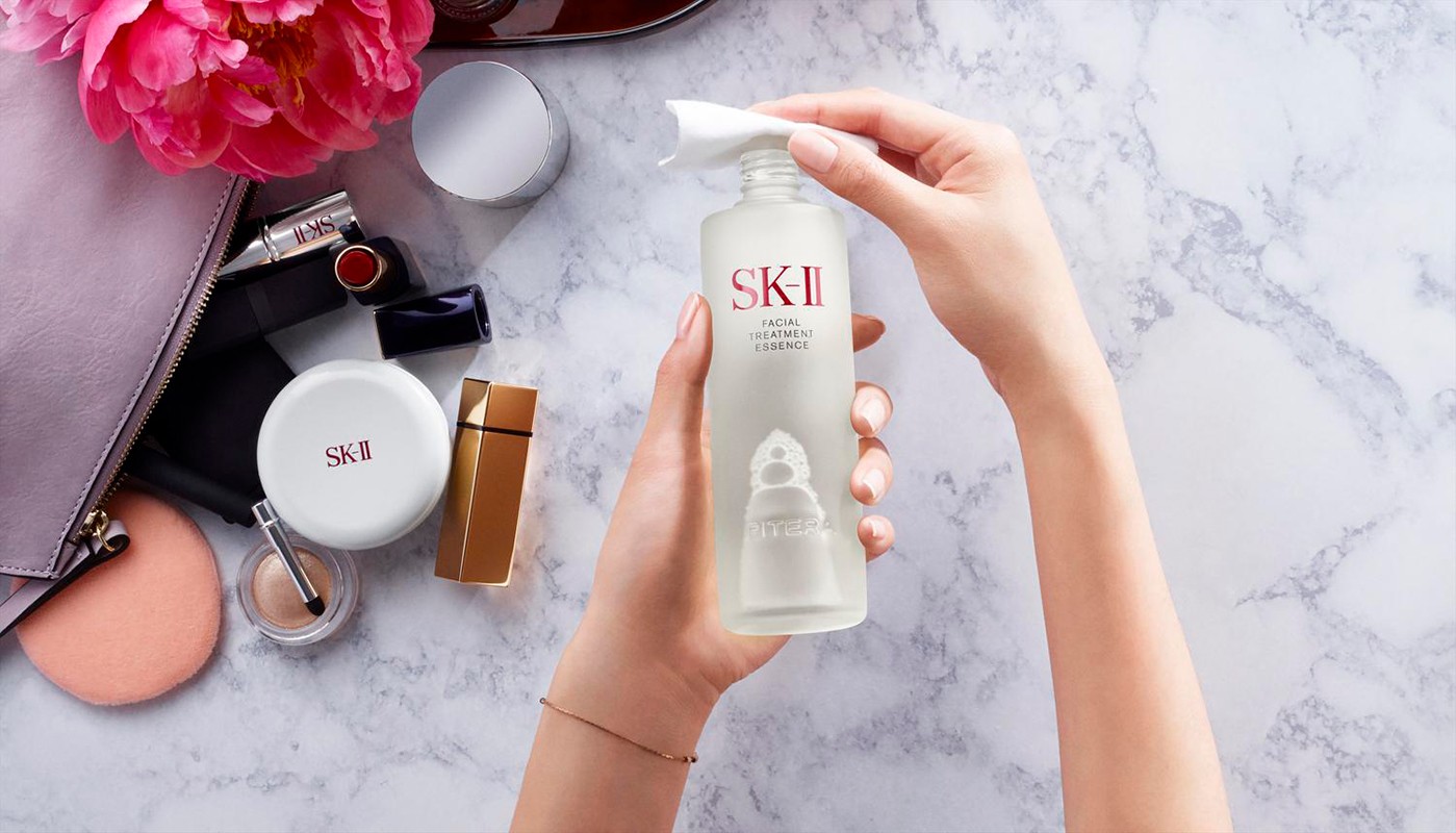 SK-II Facial Treatment Essence bottle held by hands with a tissue on top. Cosmetics and makeup bag are in the background.