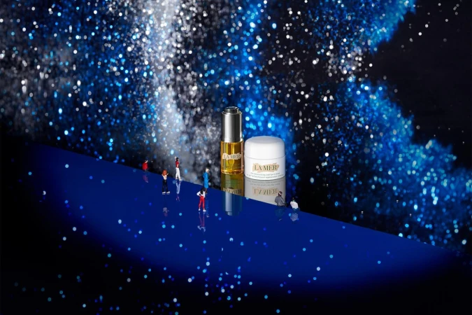 La Mer skincare products sit on a reflective blue surface with miniature figures and bokeh-lit background creating a surreal scene.