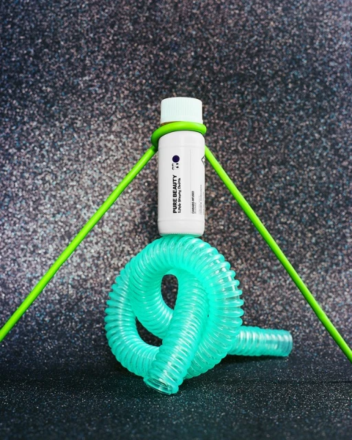 Product shot of a white bottle with green accents displayed with a green tube and blue plastic coil on a speckled background.