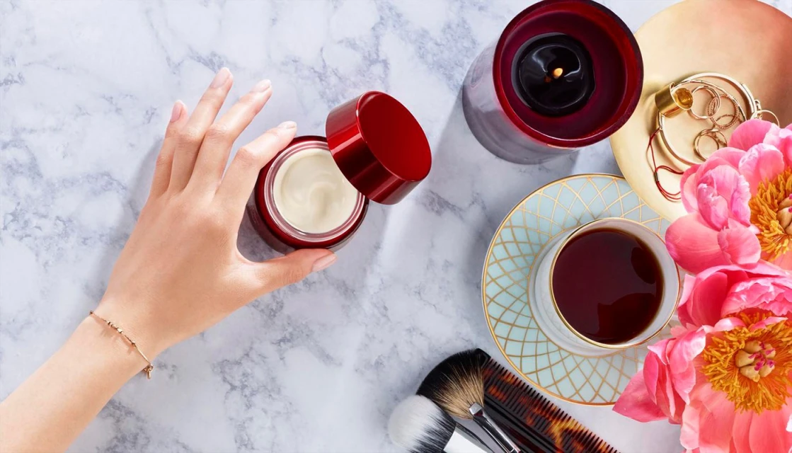 Overhead shot of a woman's hand near a cream jar, candle, cup of tea, flowers, and makeup brushes on a marble surface.