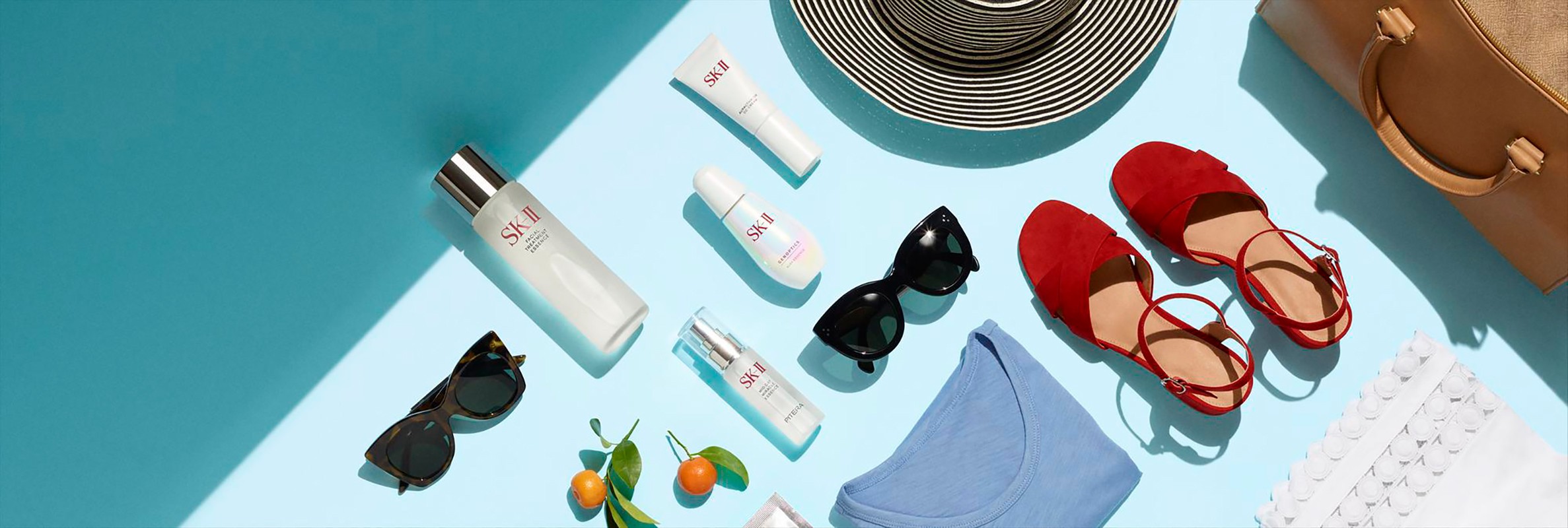 Overhead studio shot showcasing SK-II skincare products, sunglasses, red sandals, a straw hat, and a tan tote bag on a blue backdrop.