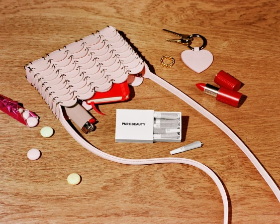 A pink scaly crossbody handbag resting on a wooden surface with a lipstick, matches, lighter, keys, and mints scattered around it.