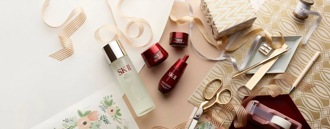 Cosmetic products including SK-II essence, cream, and eye serum arranged with gold scissors, ribbon, and gift on cream and gold surface.