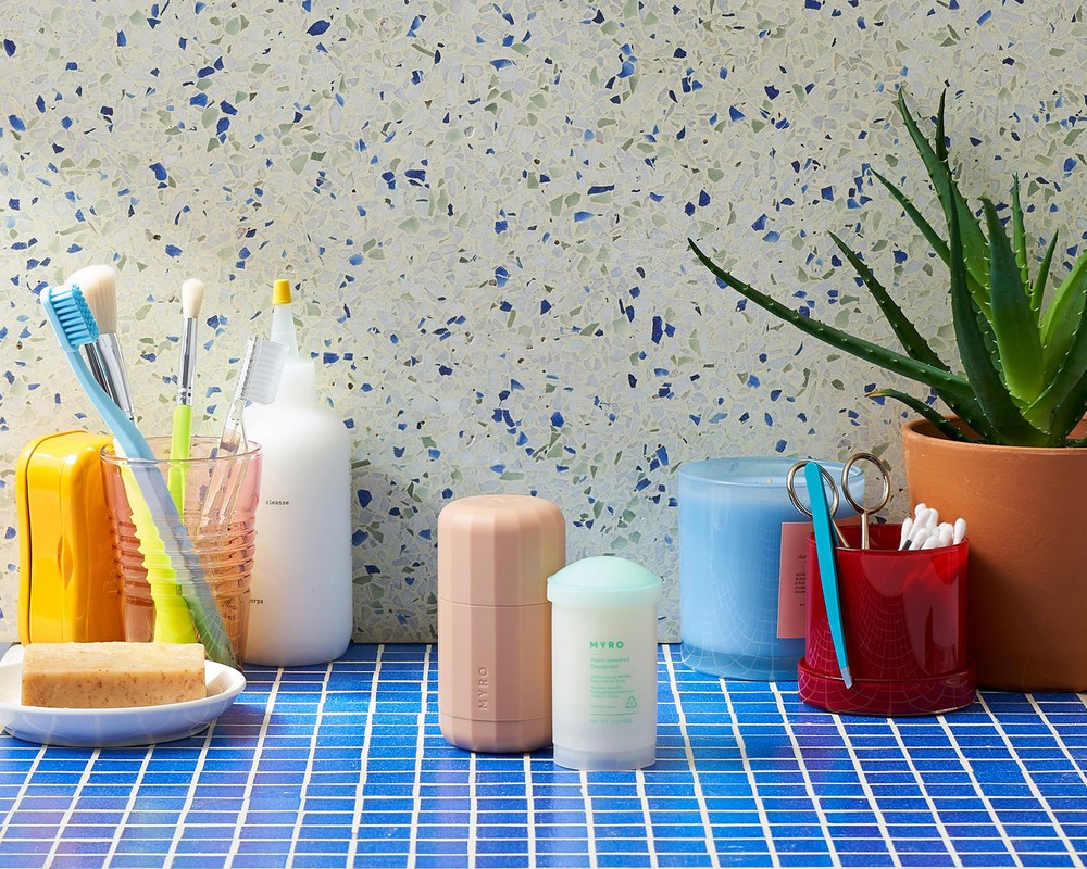 Bathroom scene featuring toiletries and personal care items on a blue tile surface with a mottled green and blue background wall.