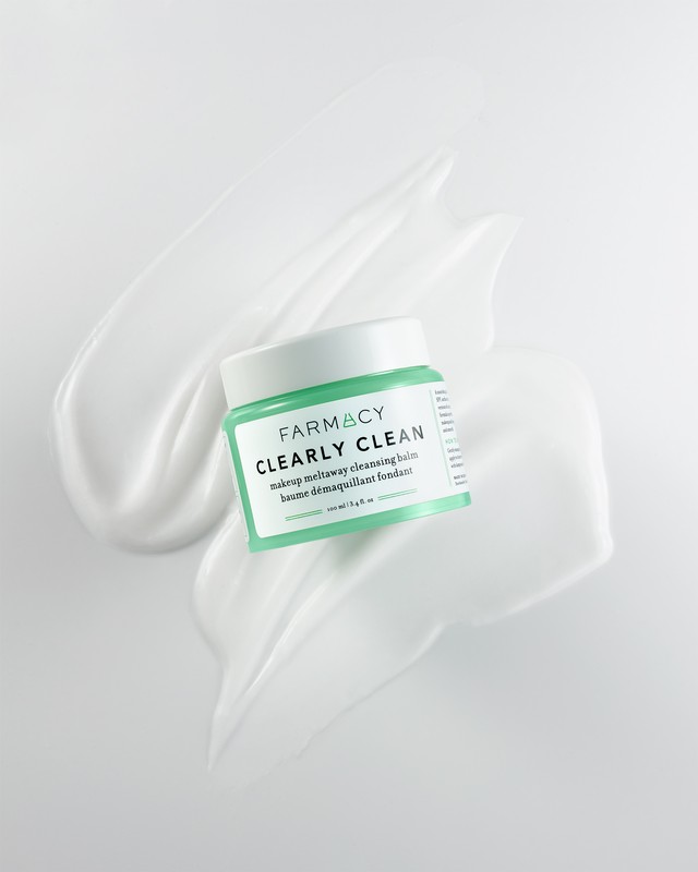 Farmacy Clearly Clean makeup removing balm with light green label, surrounded by swirls of white cream against white background.