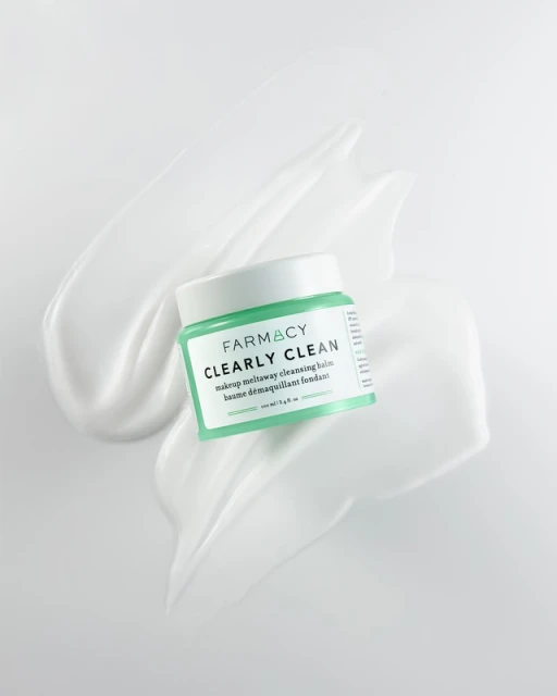 Farmacy Clearly Clean makeup removing balm with light green label, surrounded by swirls of white cream against white background.