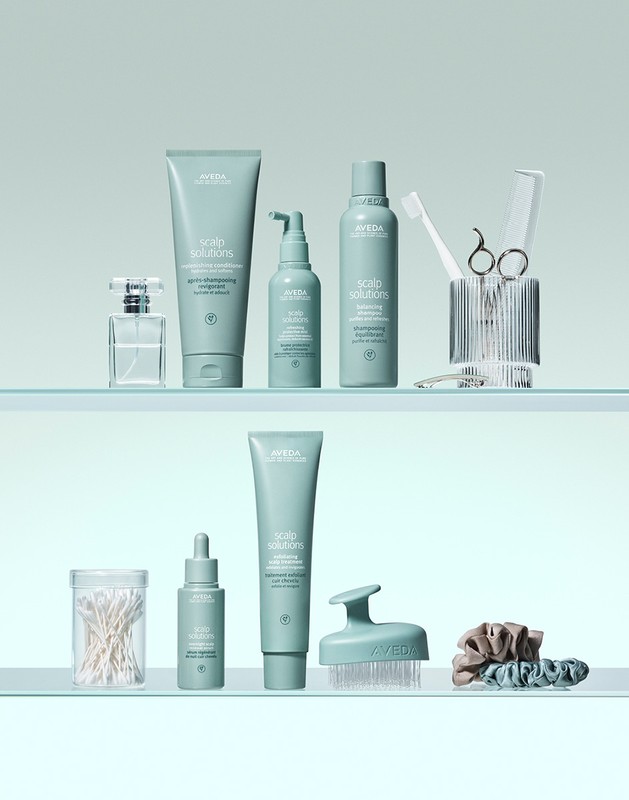 Aveda hair and scalp care products on glass shelves including shampoo, conditioner, hair oil, brush, and accessories, studio shot.