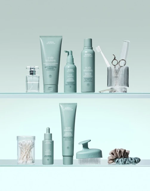 Aveda hair and scalp care products on glass shelves including shampoo, conditioner, hair oil, brush, and accessories, studio shot.